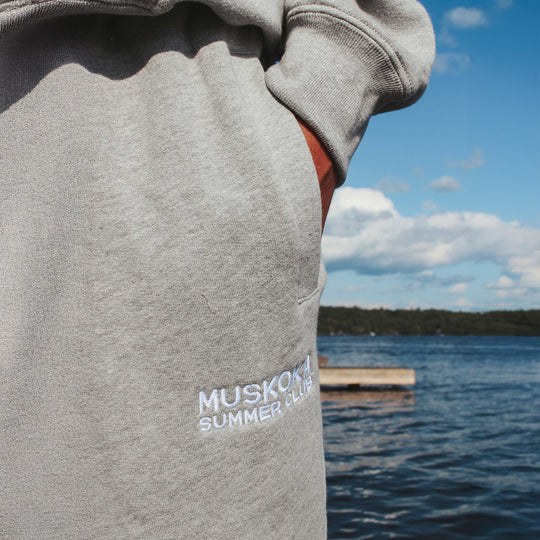Person wearing a gray sweatshirt with 'Muskoka Summer Club' text by a lake.