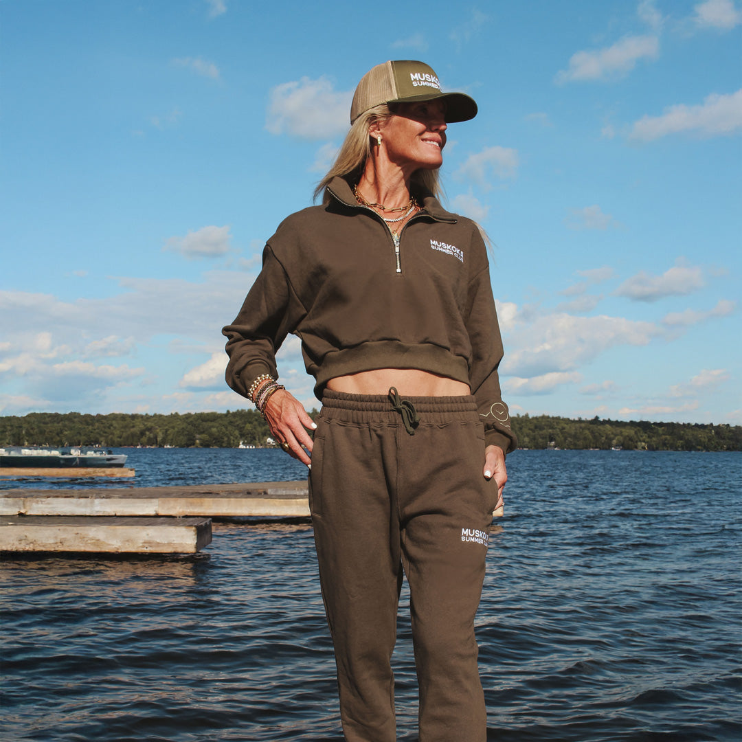 Person wearing a brown outfit with a cap standing by a lake