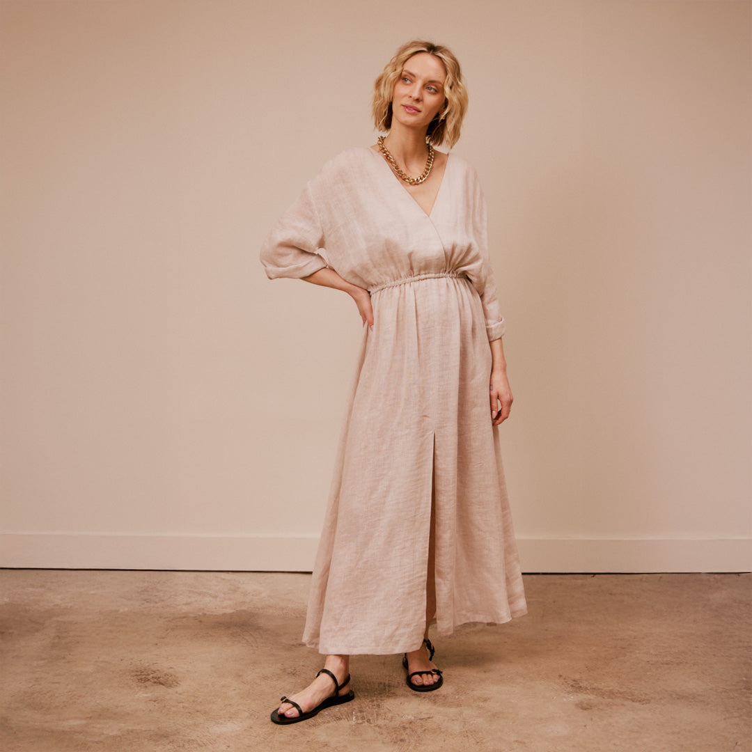 Woman wearing a long beige dress and black sandals against a plain background
