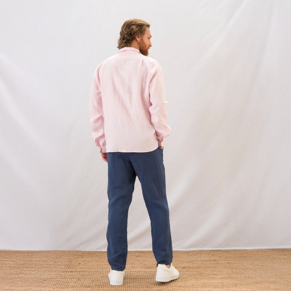 Man wearing a pink shirt and blue pants standing against a white wall.