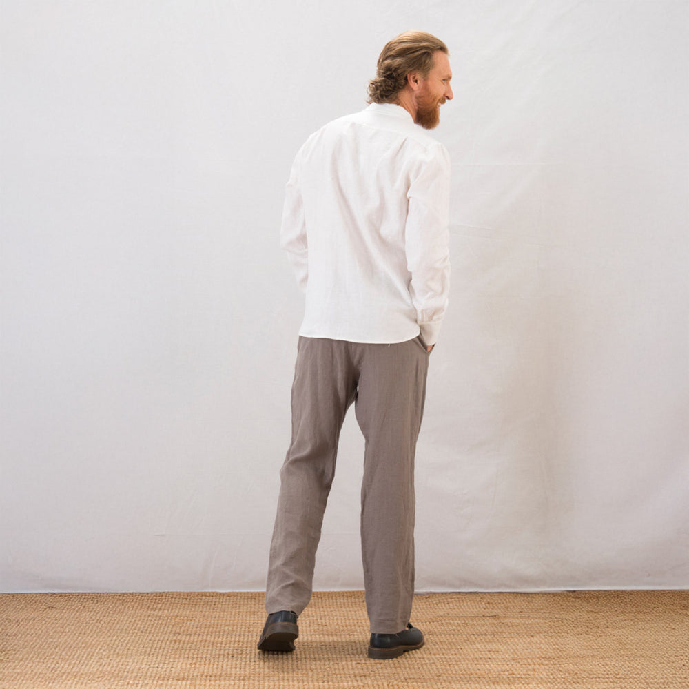 Man wearing a white shirt and gray pants standing against a plain white wall.
