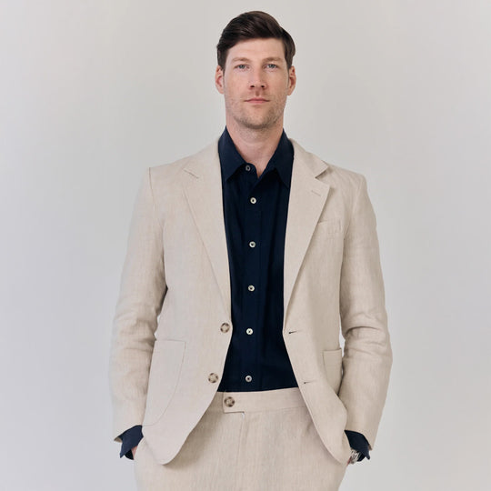 Man wearing a beige suit jacket over a navy shirt on a plain background