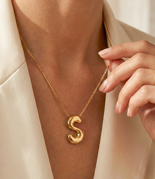 Gold necklace with a 'S' pendant on a person wearing a beige outfit.
