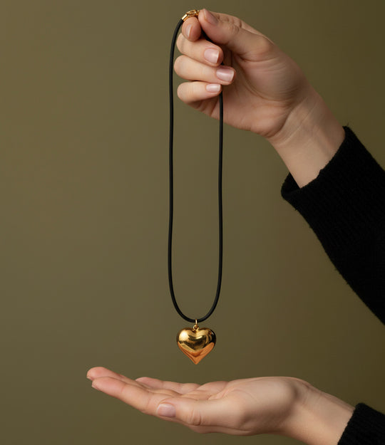 18K Gold heart-shaped pendant on a black cord held between two hands against a brown background