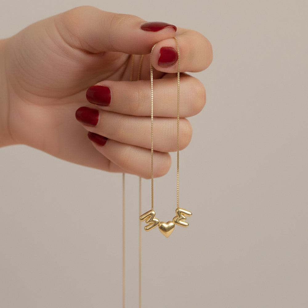 Hand holding up a delicate gold necklace, allowing the puffy gold 'M,' heart, and 'M' charms to hang.