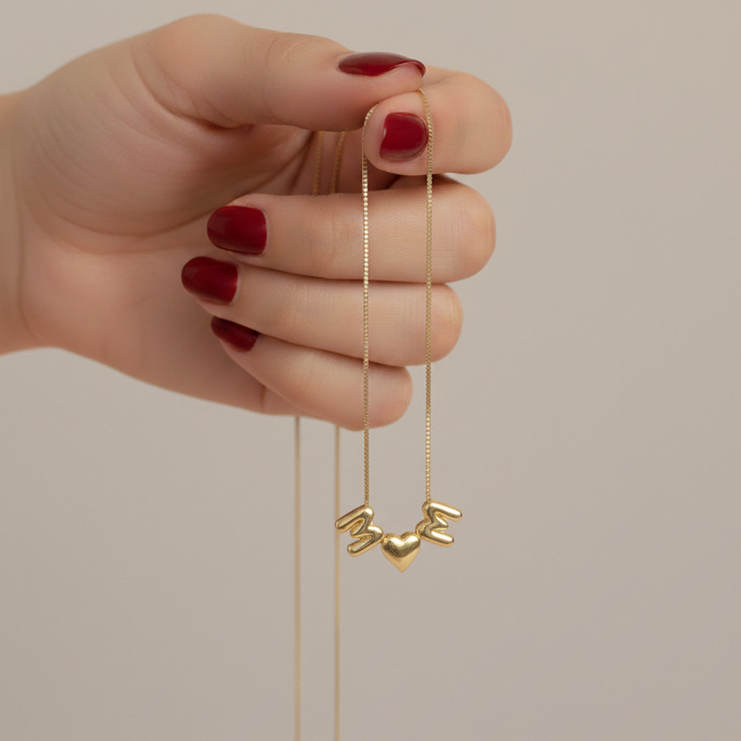 Hand holding up a delicate gold necklace, allowing the puffy gold 'M,' heart, and 'M' charms to hang.