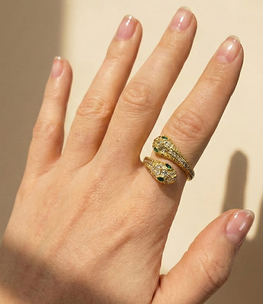 Hand wearing a gold ring with green gemstones on a beige background