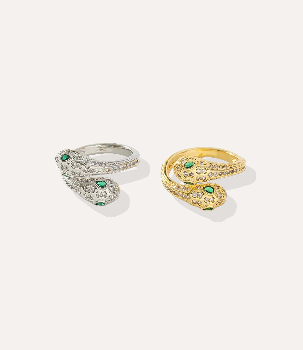 Two snake-shaped rings, one gold and one silver, with green gemstones on a light gray background.