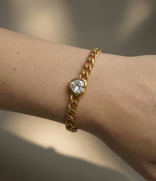 Gold chain bracelet with a heart-shaped gemstone on a wrist against a neutral background