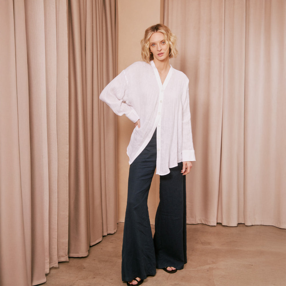 Woman wearing a white shirt and dark pants standing against a beige curtain.