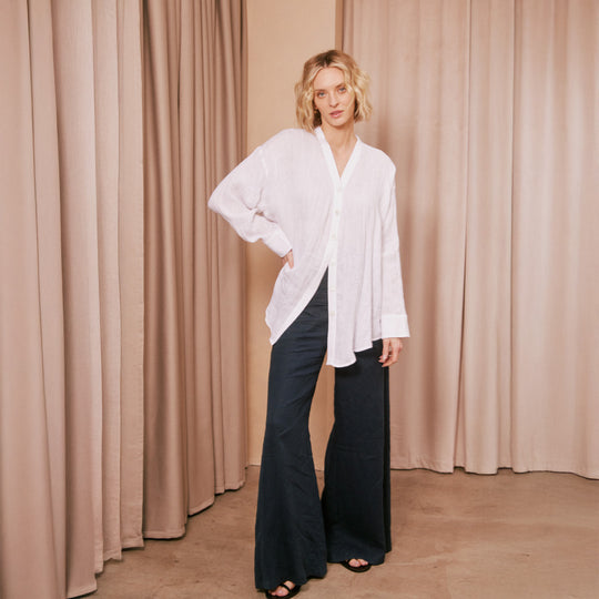 Woman wearing a white shirt and dark pants standing against a beige curtain.